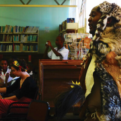 Moroka Moreri wears fur and patterned clothes, performing poetry in a room full of books