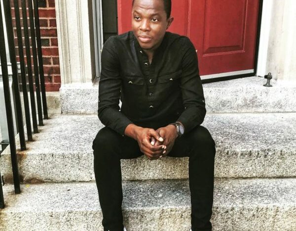 Romeo Oriogun sits outside on the front steps of a building dressed in black with white shoes